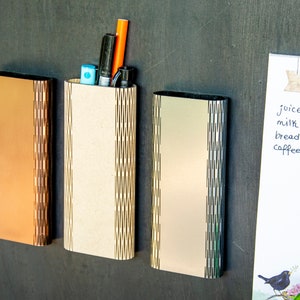 May include: Three wooden wall organisers with a decorative cut-out design. The organiser on the left is brown, the middle organiser is beige, and the organiser on the right is silver. The middle organiser has a blue pen, a black pen, and an orange pen inside.