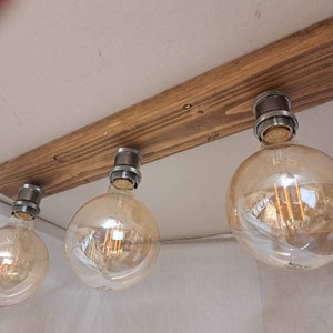 May include: Three clear glass light bulbs with warm white filament bulbs hanging from a rustic wooden beam. The beam is attached to the ceiling with metal fixtures.