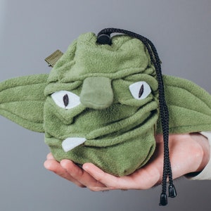 May include: A green fleece drawstring bag shaped like a character's head with large ears, white eyes, and a small nose. The bag has a black drawstring closure and a small loop on the side. The bag is held in a person's hand.
