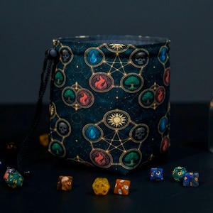 May include: A cylindrical dice bag with a drawstring closure. The bag is dark blue with a repeating pattern of skulls, trees, flames, water droplets, and suns, all connected by gold lines. Several multi-sided dice are scattered around the bag.
