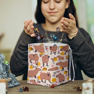 May include: A beige drawstring dice bag featuring a repeating pattern of brown capybaras, yellow circles, and pink floral accents. The bag is open, with several colorful dice being tossed into it. The bag is sitting on a wooden surface.