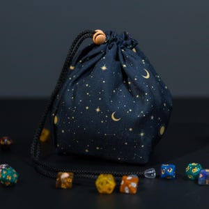 May include: A black drawstring bag with a celestial print featuring gold stars, moons, and a sun. The bag has a wooden toggle closure and a black drawstring.