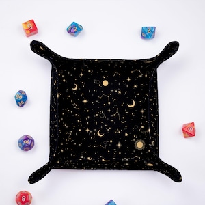 May include: Black velvet dice tray with a gold celestial print featuring moons, stars, and constellations. The tray has two fabric handles and is surrounded by colourful dice.