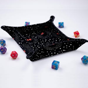 May include: Black dice tray with a repeating pattern of bats and stars. The tray is made of fabric and has a soft, plush feel. There are several colorful dice scattered around the tray.