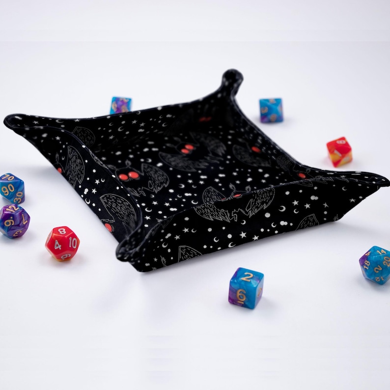 May include: Black dice tray with a repeating pattern of bats and stars. The tray is made of fabric and has a soft, plush feel. There are several colorful dice scattered around the tray.