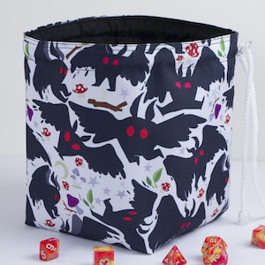 May include: A white drawstring bag featuring a repeating pattern of dark moth-like creatures with bright red eyes, interspersed with small red and purple mushrooms, green leaves, and light purple stars. The interior of the bag is lined in black fabric.