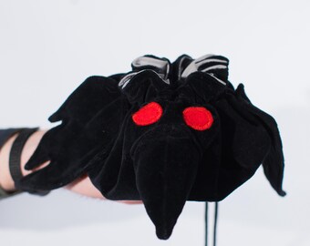May include: A black velvet drawstring bag shaped like a moth, with large red embroidered eyes. The bag has a long, pointed beak and wing-like flaps. The drawstring is black.