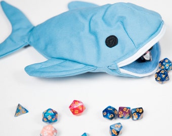May include: A light blue shark-shaped dice bag with a zipper opening. The bag is lying on a white surface with several multi-colored dice spilling out of the bag.
