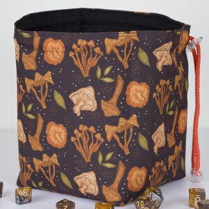 May include: A dark brown fabric drawstring bag featuring a repeating pattern of orange and tan mushrooms and small green leaves. The bag has a black interior lining and a bright orange cord with a clear plastic toggle closure for securing the top.