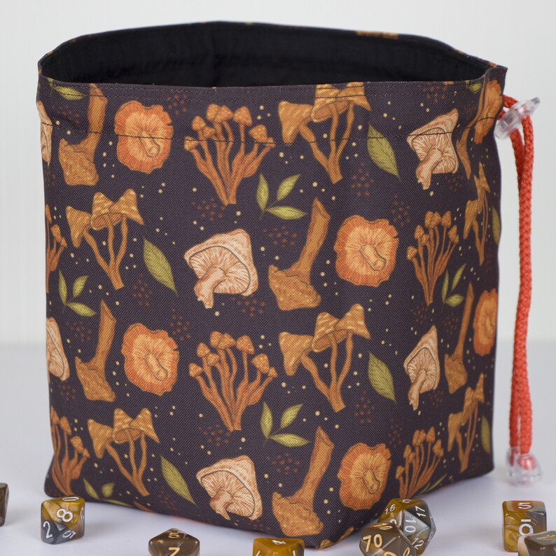 May include: A dark brown fabric drawstring bag featuring a repeating pattern of orange and tan mushrooms and small green leaves. The bag has a black interior lining and a bright orange cord with a clear plastic toggle closure for securing the top.