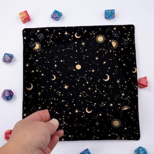 May include: A black velvet dice tray with a gold celestial pattern featuring stars, moons, and suns. The tray is folded in half, revealing a second layer of the same pattern.