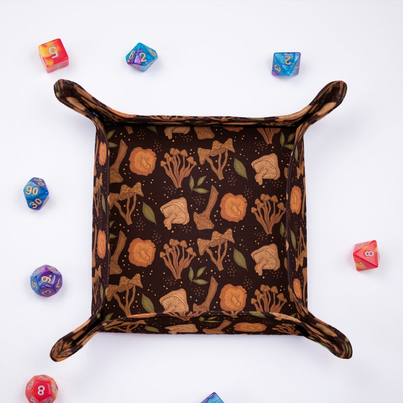 May include: A square fabric dice tray with a dark brown background featuring a repeating pattern of orange and tan mushrooms and small green leaves. The tray has four snapped corners and is surrounded by six multi-colored polyhedral dice.