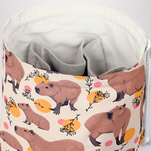 May include: A beige fabric storage bag with a repeating pattern of brown capybaras, orange and pink circles, and black floral accents. The bag has a drawstring closure and a gray interior with multiple pockets.