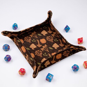 May include: A square fabric dice tray with raised corners featuring a dark brown background with a repeating pattern of tan mushrooms and small green leaves, surrounded by several multi-colored polyhedral dice with gold-colored numbers.