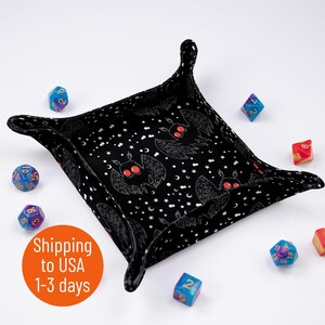 May include: Black dice tray with a repeating pattern of white moths with red eyes on a black background with white stars. The tray is made of fabric and has a square shape with rounded corners. There are several colorful dice scattered around the tray.