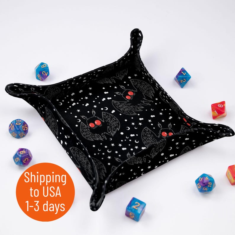 May include: Black dice tray with a repeating pattern of white moths with red eyes on a black background with white stars. The tray is made of fabric and has a square shape with rounded corners. There are several colorful dice scattered around the tray.