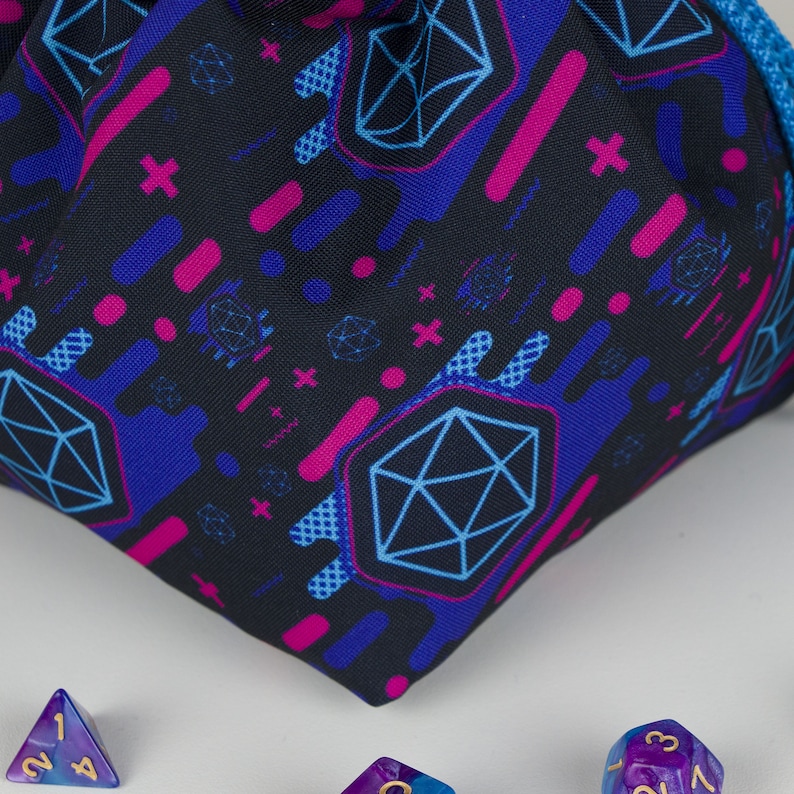 Puede incluir: Una bolsa de dados negra con un patr&oacute;n repetido de formas geom&eacute;tricas azules y magenta. La bolsa es parcialmente visible, con un cord&oacute;n azul. Varios dados poli&eacute;dricos en tonos morados y azules est&aacute;n esparcidos alrededor de la bolsa.