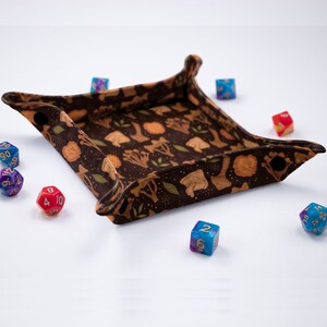 May include: A square, collapsible dice tray featuring a dark brown fabric with a repeating pattern of tan mushrooms and green leaves. The tray corners are secured with black snaps, and it is surrounded by several multi-colored polyhedral dice.