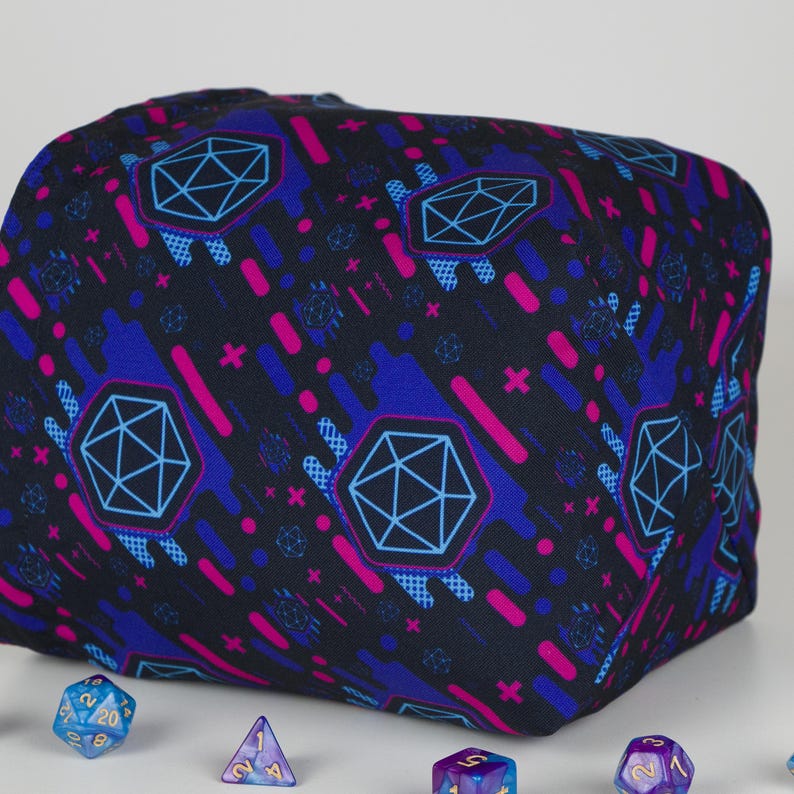 Puede incluir: Una bolsa de dados negra con un patr&oacute;n geom&eacute;trico en azul y rosa. El patr&oacute;n incluye formas de diamante, signos m&aacute;s y l&iacute;neas diagonales. Varios dados de m&uacute;ltiples lados son visibles en primer plano, en tonos de azul, morado y dorado.