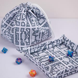 May include: A white fabric drawstring bag and a matching square folding dice tray, both featuring a black dungeon map print. Four multi-colored polyhedral dice are scattered nearby, including blue, purple, and orange translucent designs.