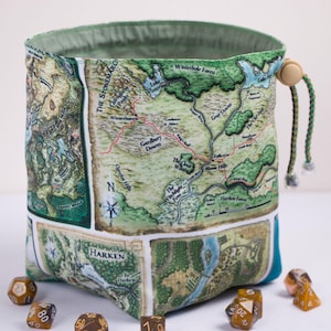 May include: A cylindrical fabric drawstring bag featuring a printed fantasy map with labels such as The Stonemarch, Winterbole Forest, and Fallcrest. The bag has a light green interior lining, a beige toggle, and braided cords, surrounded by several polyhedral dice.