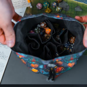 Peut inclure: Un sac à cordon noir avec un motif coloré de différentes créatures et objets fantastiques. Le sac est ouvert et rempli de plusieurs dés de différentes couleurs et numéros.
