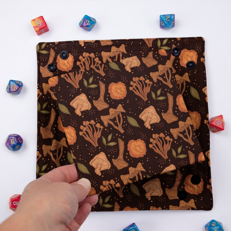 May include: A square fabric dice tray in dark brown featuring a repeating pattern of tan mushrooms and small green leaves. The tray has black snap fasteners at each corner to create raised edges, surrounded by several multi-colored polyhedral dice.