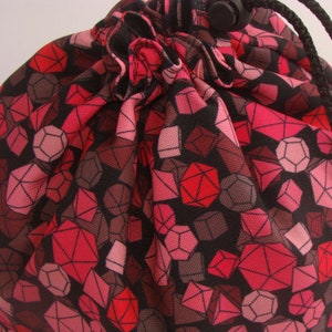 May include: A drawstring bag with a repeating geometric dice pattern in shades of red, pink, and brown on a black background. The bag is gathered at the top with a black drawstring and a black plastic toggle.