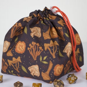 May include: A dark brown fabric drawstring pouch featuring a repeating pattern of orange and tan mushrooms with small green leaves and gold dots. The bag is cinched at the top with a bright orange cord and a clear plastic toggle, surrounded by dice.