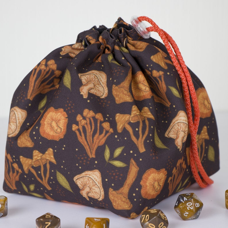 May include: A dark brown fabric drawstring pouch featuring a repeating pattern of orange and tan mushrooms with small green leaves and gold dots. The bag is cinched at the top with a bright orange cord and a clear plastic toggle, surrounded by dice.
