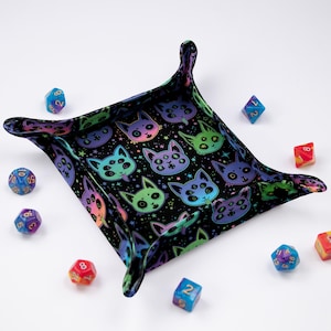May include: A black, square dice tray with a colorful cat face pattern. The tray has raised edges and is surrounded by various colorful dice. The cat faces are in shades of green, blue, purple, and pink.
