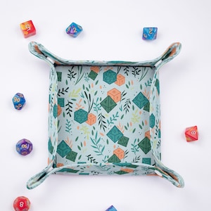 May include: A square fabric dice tray with a light blue background featuring a pattern of green leaves, orange sprigs, and various polyhedral dice. The tray has folded corners secured with fasteners and is surrounded by several loose multi-coloured dice.