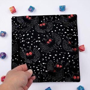 May include: Black dice bag with a white and red moth pattern. The bag is made of fabric and has a drawstring closure. The bag is filled with dice.