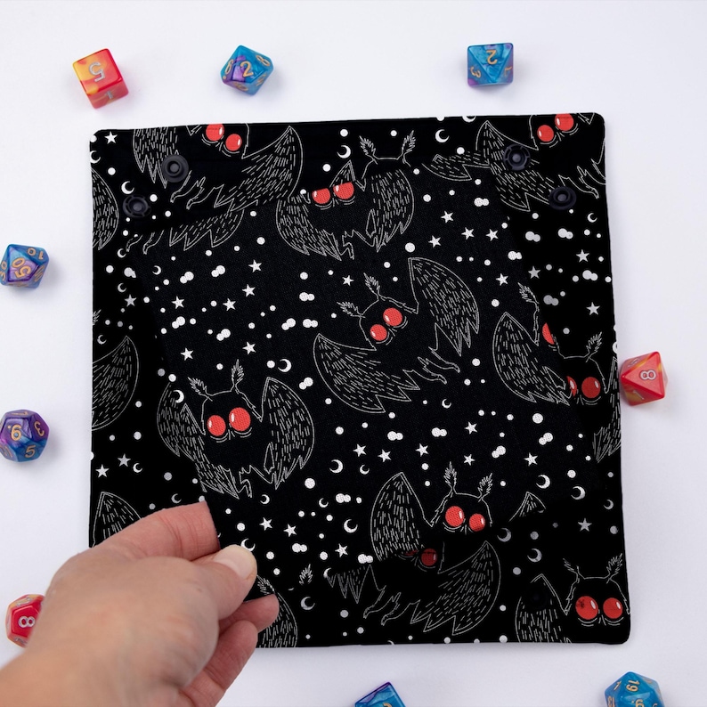 May include: Black dice bag with a white and red moth pattern. The bag is made of fabric and has a drawstring closure. The bag is filled with dice.