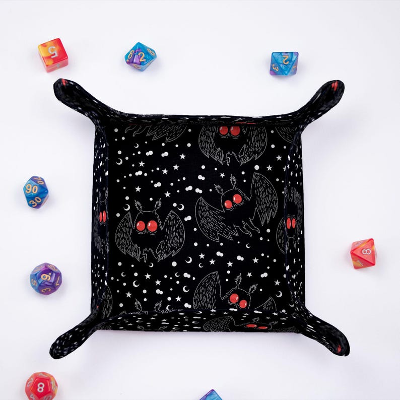 May include: Black dice tray with a repeating pattern of white moths with red eyes on a black background with white stars and crescent moons. The tray has a black felt lining and four black fabric flaps.