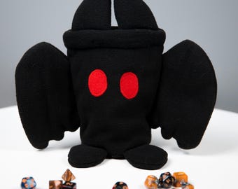 May include: A black plush creature with red eyes and wings, resembling a bat, sits on a white surface. Assorted polyhedral dice in various colors are scattered around the plush toy, suggesting a tabletop gaming theme.