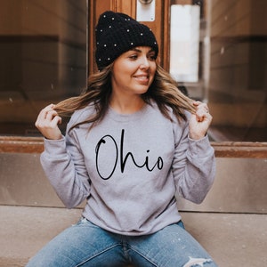 May include: A woman wearing a gray sweatshirt with the word "Ohio" written in black script. She is wearing a black beanie with white dots and blue jeans.