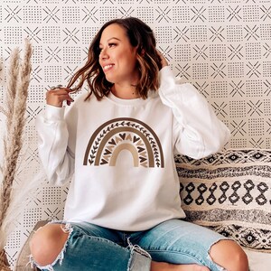 May include: A white sweatshirt with a brown and beige rainbow graphic featuring hearts and leaves. The rainbow is centered on the front of the sweatshirt.