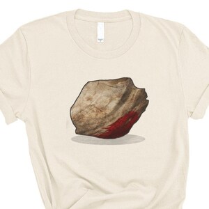 May include: A beige t-shirt with a graphic of a large, gray rock with a red stain on it.