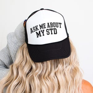 May include: A black and white trucker hat featuring a foam front panel with the text ASK ME ABOUT MY STD printed in a bold black collegiate font. The cap includes a black brim and a mesh back panel with an adjustable snapback closure.