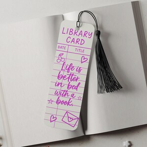 May include: A white bookmark with a black tassel, featuring the text "LIBRARY CARD" and "Life is better in bed with a book" in purple. The bookmark has a library card design with decorative hearts, cherries, and stars.