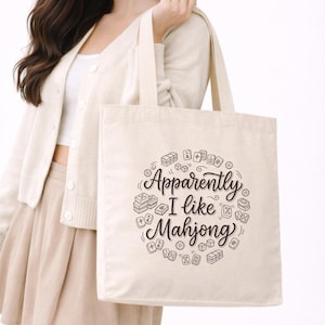 Puede incluir: Un bolso de tela beige con un estampado gráfico negro de varias fichas de Mahjong dispuestas en un patrón circular alrededor del texto "Apparently I like Mahjong" escrito con una fuente cursiva decorativa en el centro.
