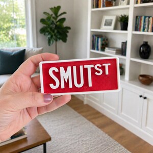 Puede incluir: Un letrero rectangular con borde blanco y fondo rojo que presenta el texto blanco en relieve SMUTST. El letrero tiene un estilo de fuente de letras de bloque y un acabado liso, presentando una visualizaci&oacute;n clara y legible de las letras.