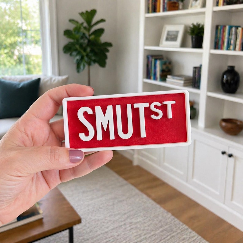 Puede incluir: Un letrero rectangular con borde blanco y fondo rojo que presenta el texto blanco en relieve SMUTST. El letrero tiene un estilo de fuente de letras de bloque y un acabado liso, presentando una visualizaci&oacute;n clara y legible de las letras.