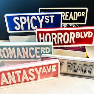 Puede incluir: Una colecci&oacute;n de letreros rectangulares impresos en 3D con letras blancas en relieve. Los letreros incluyen textos como SPICY ST, READ DR, HORROR BLVD, ROMANCE RD, FANTASY AVE y MY READS en varios colores como azul, negro, rojo, azul claro, rosa y gris.