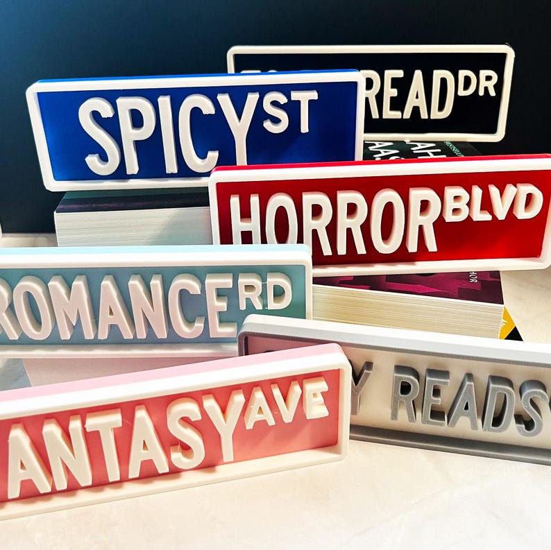 Puede incluir: Una colecci&oacute;n de letreros rectangulares impresos en 3D con letras blancas en relieve. Los letreros incluyen textos como SPICY ST, READ DR, HORROR BLVD, ROMANCE RD, FANTASY AVE y MY READS en varios colores como azul, negro, rojo, azul claro, rosa y gris.