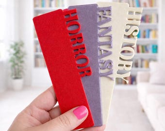 Könnte beinhalten: Drei rechteckige Lesezeichen in Rot, Lila und Creme, die jeweils vertikale Ausschnittbuchstaben aufweisen. Das rote Lesezeichen zeigt das Wort HORROR, das lila Lesezeichen liest FANTASY und das cremefarbene zeigt das Wort HISTORY.