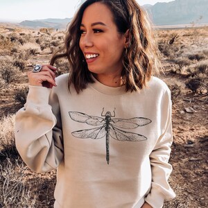 May include: A beige crewneck sweatshirt featuring a detailed black line art illustration of a dragonfly centered on the chest. The garment has long sleeves with ribbed cuffs and a ribbed neckline, constructed from a solid-colored fabric.