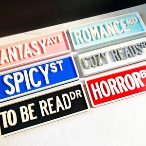 Puede incluir: Una colecci&oacute;n de seis letreros rectangulares con letras blancas en relieve. Los letreros muestran el texto FANTASY AVE, ROMANCE RD, COZY READS RD, SPICY ST, TO BE READ DR y HORROR BLVD, cada uno sobre un fondo de diferente color, incluyendo rosa, azul, gris y rojo.
