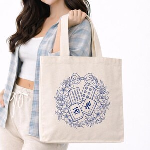 Puede incluir: Un bolso de tela beige con una ilustración de líneas azules de tres fichas de mahjong rodeadas por una corona floral y un lazo. Las fichas muestran los caracteres para Oeste y Este junto con patrones de bambú y puntos en un diseño circular.